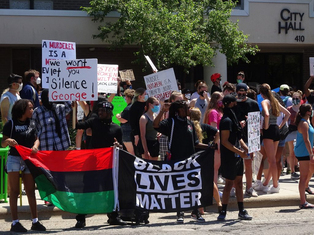Demonstrators at a Black Lives Matter protest in East Lansing, Michigan.  Protesters in the foreground carry an African flag and a Black Lives Matter flag in front of them.  Other protesters carry signs.  Only one sign is legible, and it reads "we wont let you silence George."  The four protesters in the foreground are African American women wearing black.  The protesters behind them are a mix of races.  