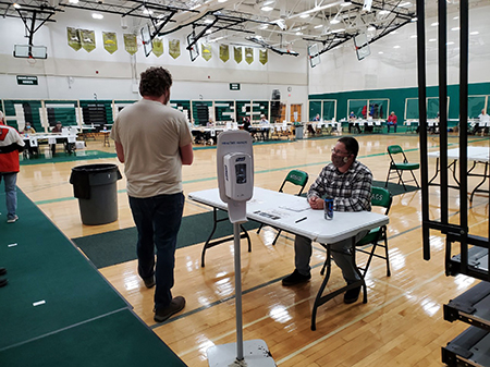 A voter had to be a really engaged citizen to vote in the recent Wisconsin primary, in the midst of the pandemic.  This image shows such an engaged citizen checking with National Guard member serving as a poll worker in what appears to be a high school gym.  