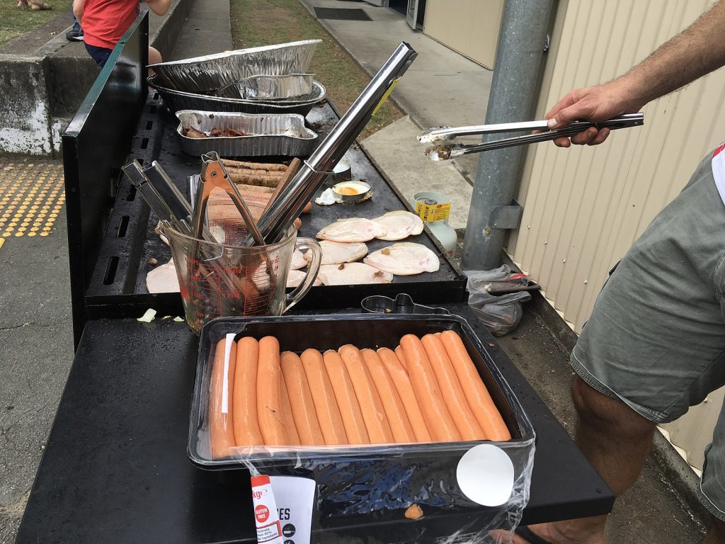 A container of uncooked hotdogs is in the foreground.  Further back are some kitchen implements in a measuring cup, and a long grill with bread toasting, hot dogs cooking, and some tinfoil pans.  A disembodied male hand wields tongs, and a disembodied leg is in frame wearing shorts.  Democracy Sausages are an important tradition of Australian voting.  