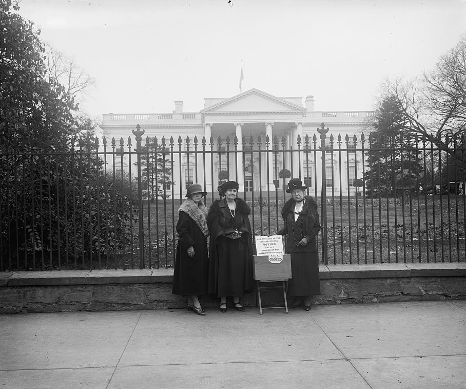 A black and white image of three League of Women Voters members in long coats, fancy hats, and jewelry, standing outside the White House with a display that is too small to read.  The League of Women Voters has been putting out Voter Guides for a long time.  